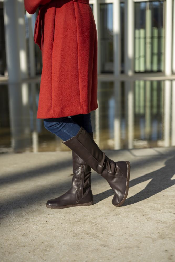 Eine Person in einem roten Mantel und braunen kniehohen ZAQQ QATCH Winter Brown-Stiefeln geht auf einem sonnenbeschienenen Gehweg. Im Hintergrund ist ein modernes Glasgebäude mit reflektierenden Oberflächen zu sehen.