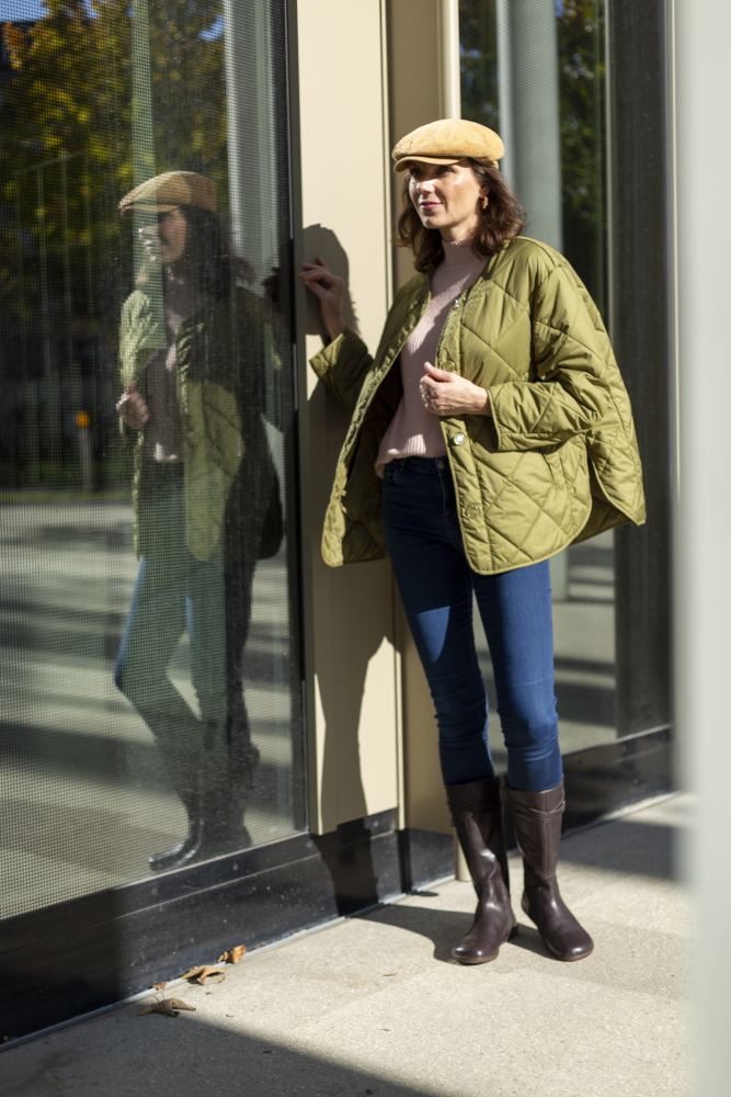 Eine Frau mit beigem Hut, grüner Steppjacke, rosa Pullover und Bluejeans steht vor einer Glaswand. Im Freien schimmert ihr Spiegelbild im Glas, während sie selbstbewusst die Wand berührt und die ATTRAQT Nappa Brown Boots von ZAQQ trägt.