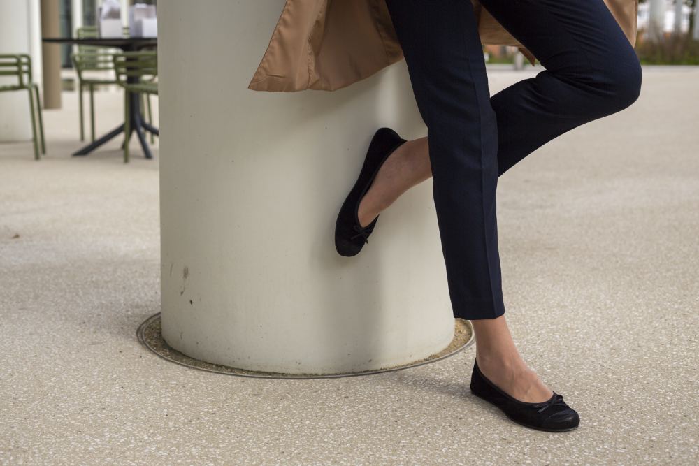 Eine Person in dunkelblauer Hose und Schuhen aus der TWIST Velours Black Kollektion von ZAQQ lehnt an einer großen weißen Säule. Im Hintergrund sind Tische und Stühle auf einer gepflasterten Fläche zu sehen, die eine Atmosphäre von Komfort und Flexibilität schaffen.