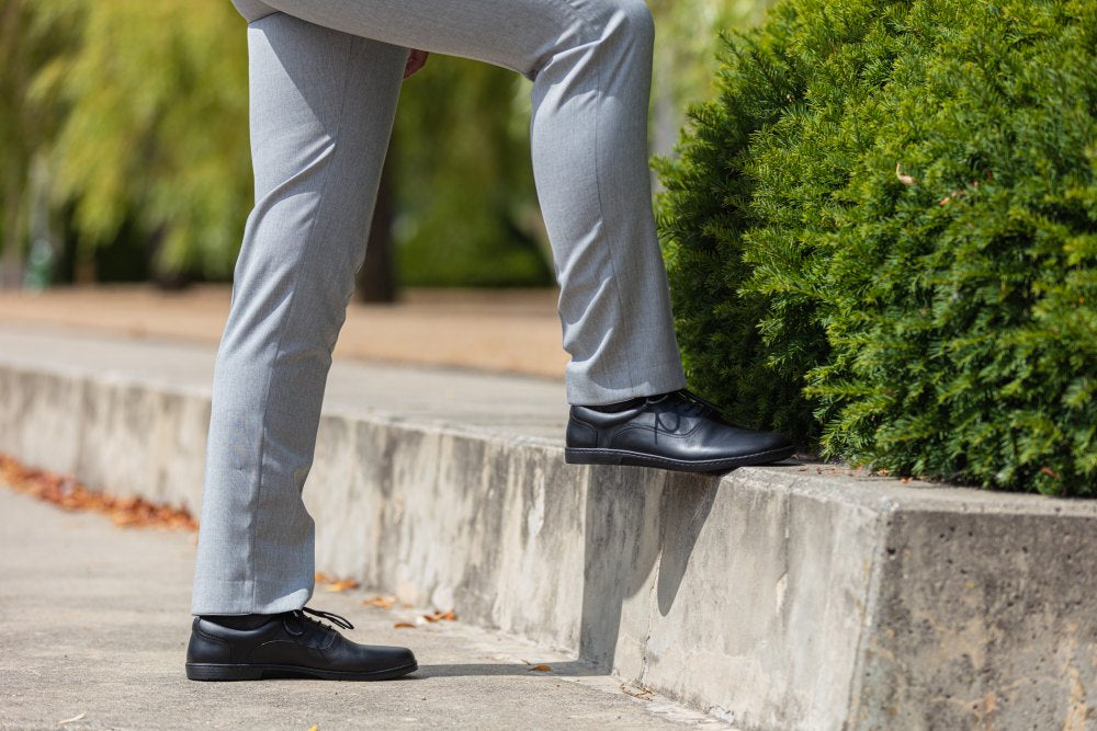 Eine Person in grauer Anzughose und einem Paar schwarzer ZAQQ APEQ-Schuhe tritt auf einen Betonvorsprung, im Hintergrund bilden grüne Sträucher und Bäume eine natürliche Kulisse.