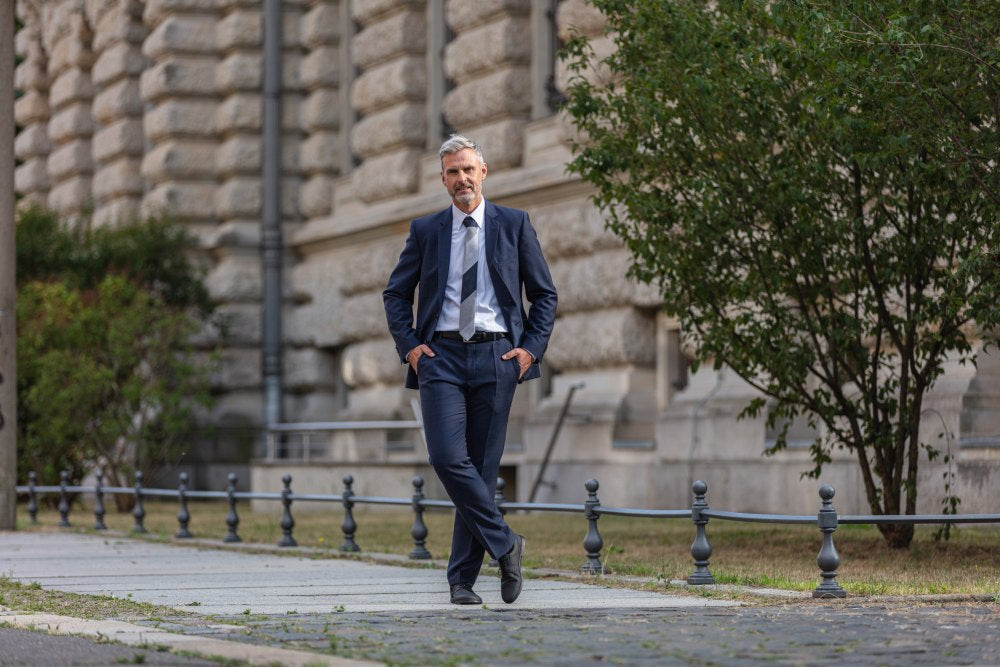 Ein Mann im marineblauen Anzug steht selbstbewusst mit den Händen in den Taschen auf einem Kopfsteinpflasterweg. Mit den eleganten BRIQ Brogue Vegan Black-Schuhen von ZAQQ strahlt er Stil und Eleganz aus. Vor einem historischen Gebäude mit strukturierter Steinfassade steht auch ein grüner Baum in der Nähe.