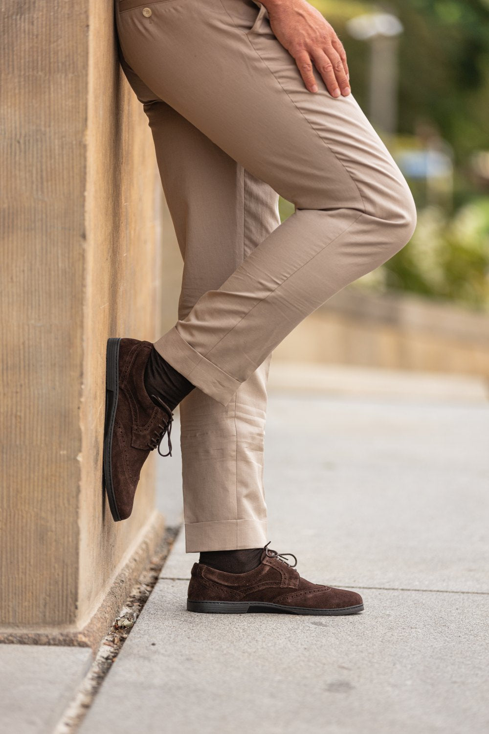 Eine Person lehnt im Freien an einer Wand und trägt beigefarbene Hosen und BRIQ Brogue Velours Brown-Schuhe von ZAQQ. Diese braunen Herrenschuhe aus Wildleder aus hochwertigem französischem Kalbleder ergänzen das lässige Ambiente perfekt und lenken die Aufmerksamkeit auf die stilvolle untere Hälfte des Outfits.