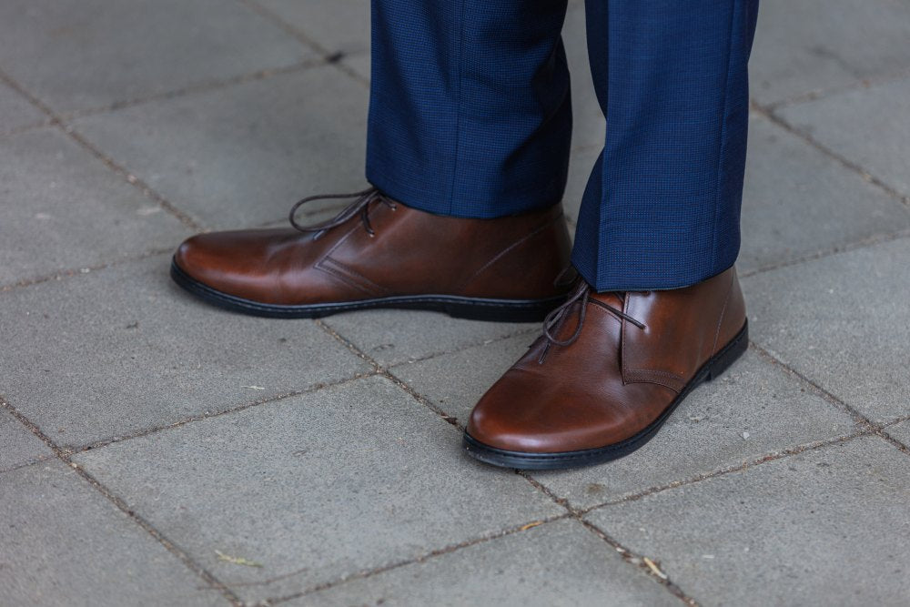 Eine Person, die DUQE Antique Brown Lederschuhe und eine blaue Hose von ZAQQ trägt, steht auf einem grau gefliesten Boden. Die älteren Halbstiefel sind poliert, sauber geschnürt und aus atmungsaktivem Glattleder gefertigt.