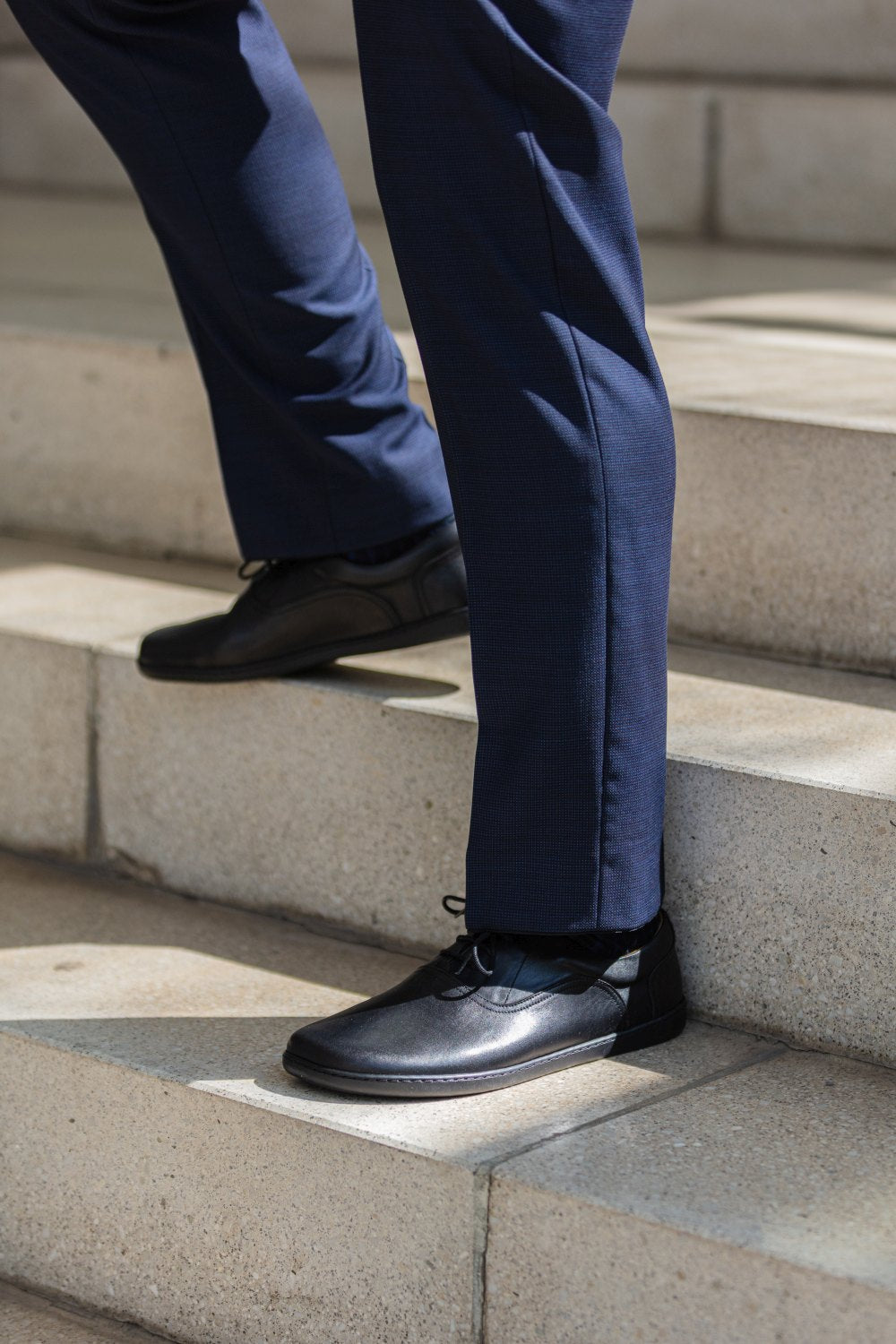 Eine Person in marineblauen Hosen und eleganten schwarzen SEQUENT-Schuhen von ZAQQ geht eine Steintreppe im Freien hinauf, wobei der Fokus auf ihren Beinen und Füßen liegt.