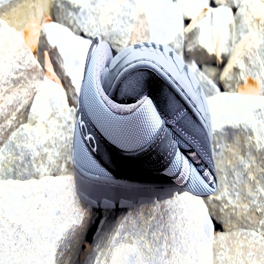 Ein SEEQ Black Kletterschuh von ZAQQ liegt seitlich auf einem Steinhaufen. Dieser Schuh aus strapazierfähigem Meshgewebe verfügt über eine robuste Sohle und einen Klettverschluss und ist somit perfekt für Kletterabenteuer im Freien.