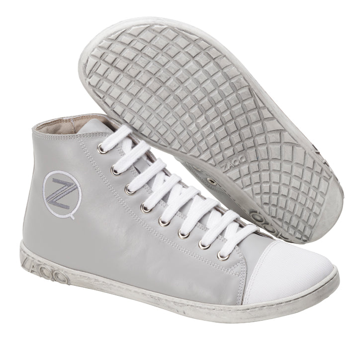 Ausgestellt sind die CHUQQS Grey High-Top-Sneaker von ZAQQ aus Nappaleder. Ein Schuh liegt auf der Seite und zeigt ein strukturiertes Sohlenmuster, das an einen Barfußschuh erinnert, während der andere aufrecht steht und Schnürsenkel und ein rundes Logo mit dem Buchstaben Z zeigt.