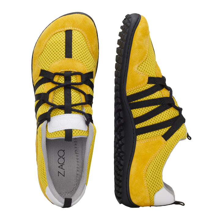 Erleben Sie die stylischen Barfußschuhe RIQ Yellow von ZAQQ. Dieses Paar leuchtend gelber Sneaker verfügt über schwarze Akzente, wobei der linke Schuh von oben gezeigt wird, um seine Mesh- und Wildledertexturen zu präsentieren. Der rechte Schuh wird in einer Seitenansicht präsentiert und zeigt das dicke Profil und das Schnürmuster für maximale Attraktivität und ein flexibles Tragegefühl.