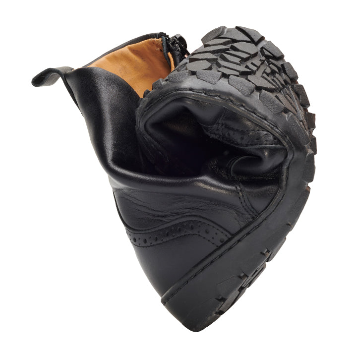 Ein einzelner QUINTIC Brogue Black-Stiefel von ZAQQ ist am Knöchel in der Mitte gebogen, was seine Flexibilität unterstreicht. Die robuste Sohle, die an einen Barfußschuh erinnert, zeigt nach oben und zeigt ihr Profilmuster und das sichtbare Innenfutter, alles vor einem schlichten weißen Hintergrund.