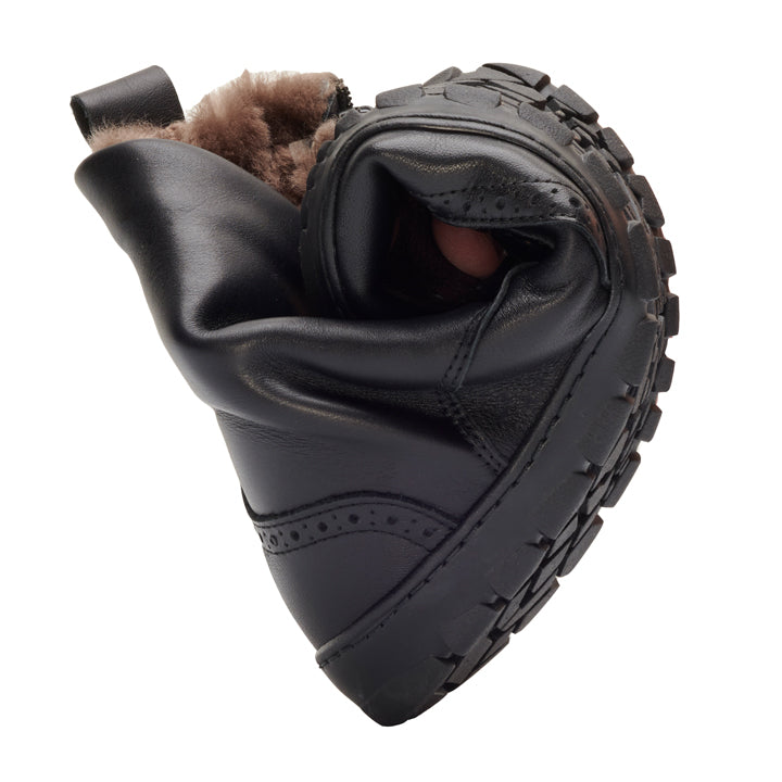 Ein flexibler QUINTIC Winter Brogue Black von ZAQQ mit robuster Sohle und Fellfutter ist gedreht, um seine Biegsamkeit zu demonstrieren. Der aus hochwertigem Nappaleder gefertigte Schuh ist mit dem Fell nach oben positioniert und die Sohle zeigt ihr strukturiertes Griffmuster.