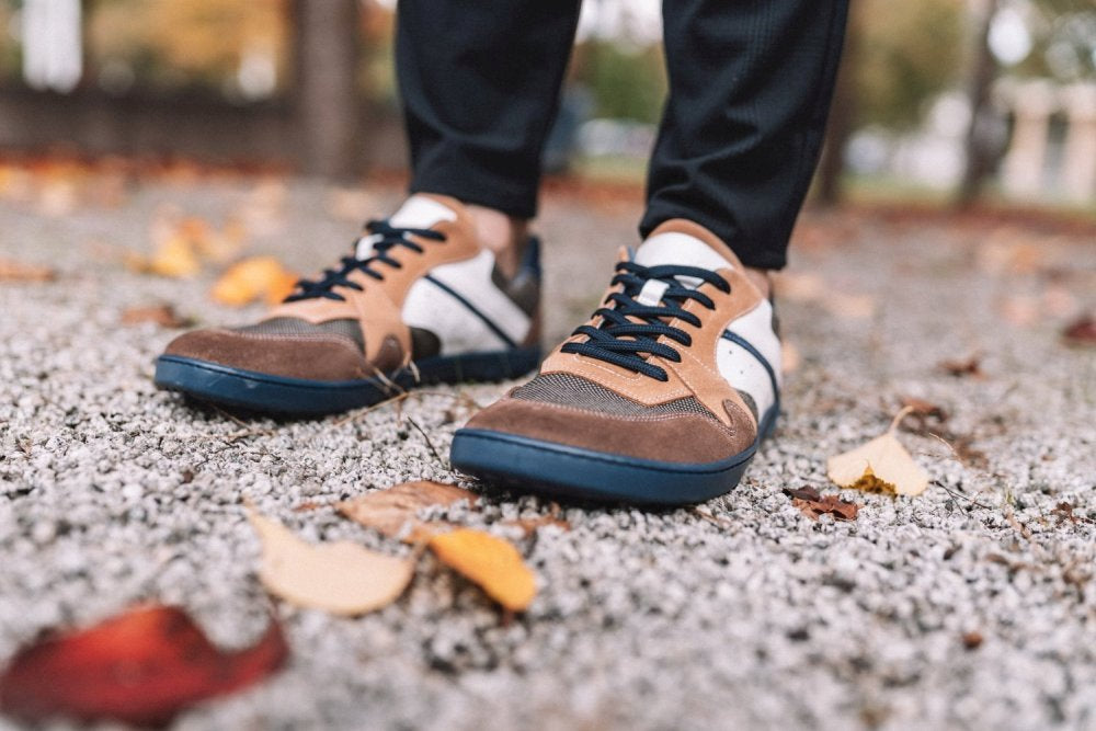 Eine Nahaufnahme zeigt jemanden, der die BREAQ Sierra-Sneaker von ZAQQ in Braun und Weiß mit schwarzen Schnürsenkeln trägt, während er auf einem mit Herbstlaub übersäten Kiesweg steht. Das schlichte Design beinhaltet die flexible ZAQQ-Sohle für außergewöhnlichen Komfort. Sie tragen schwarze Hosen, während verschwommene Bäume den Hintergrund füllen.