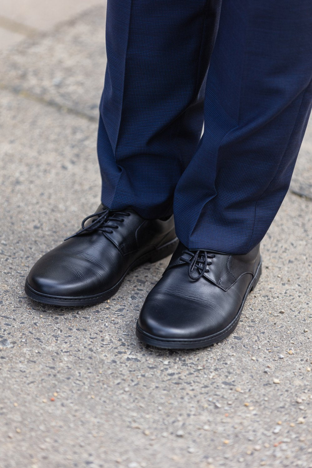 Nahaufnahme einer Person, die schwarze ZAQQ BRIQ-Schuhe und marineblaue Anzughosen trägt und auf einer Betonfläche steht und ein zeitloses Design zur Schau stellt.