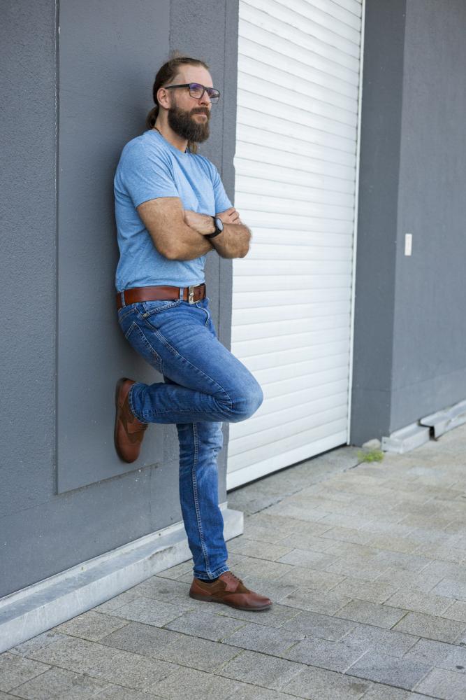 Ein Mann mit Bart und Brille lehnt mit verschränkten Armen an einer grauen Wand. Er trägt ein blaues T-Shirt, Jeans und braune BRIQ Brogue Antique Cognac-Schuhe von ZAQQ und steht vor einem weißen Garagentor auf einem gefliesten Bürgersteig.