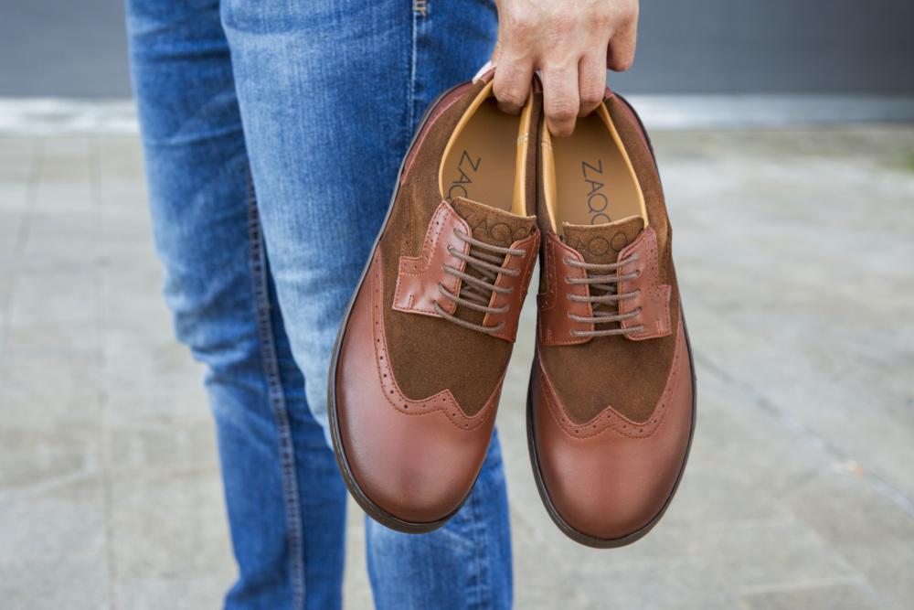 Eine Person in Jeans hält ein Paar BRIQ Brogue Antique Cognac-Schuhe an den Schnürsenkeln. Diese raffinierten Schuhe von ZAQQ zeichnen sich durch eine Kombination aus matten und glänzenden Oberflächen aus und heben sich vor einem verschwommenen urbanen Hintergrund ab.