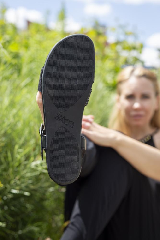 In Schwarz gekleidet präsentiert eine Person eine feminine Sandale aus der BROOQE Black-Kollektion der Marke ZAQQ, deren Sohle mit dem ZAQQ-Logo geprägt ist. Der Hintergrund aus grünem Gras und blauem Himmel akzentuiert die Verarbeitung aus Nappa- und Veloursleder wunderschön.