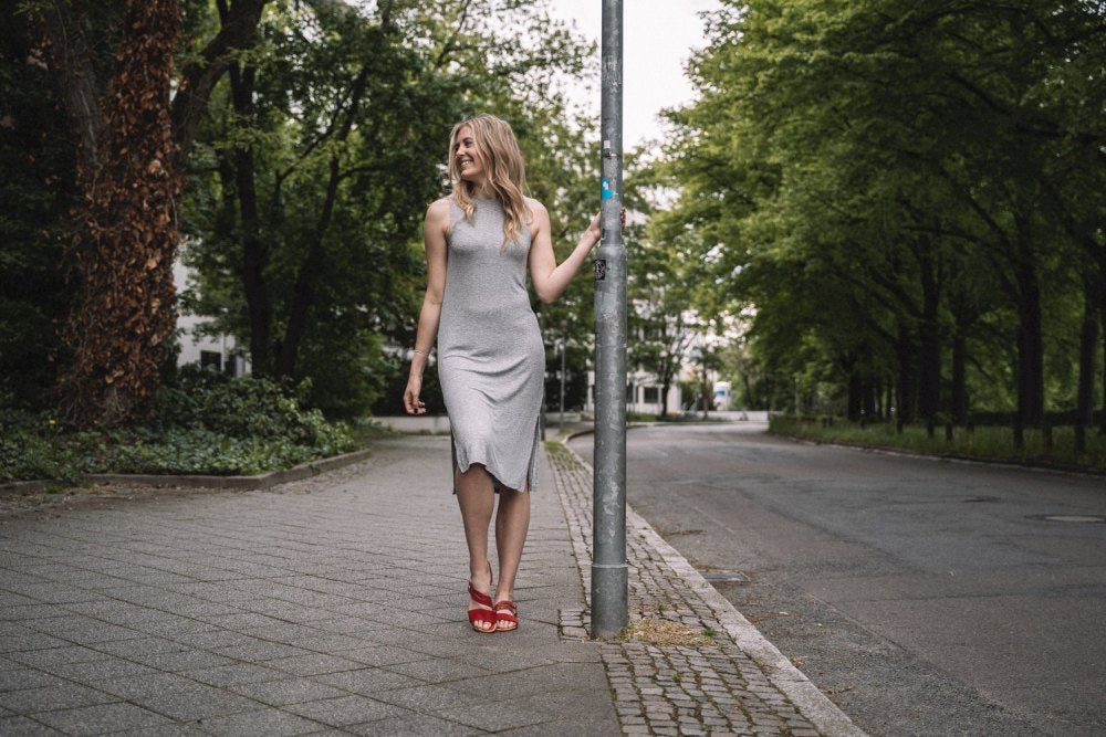 Eine Frau in einem grauen Kleid und roten ZAQQ BROOQE Sandalen steht neben einem Laternenpfahl in einer ruhigen, von Bäumen gesäumten Straße. Die Szene ist entspannt, und das grüne Laub verstärkt das Gefühl von Tragekomfort in der ruhigen Umgebung.