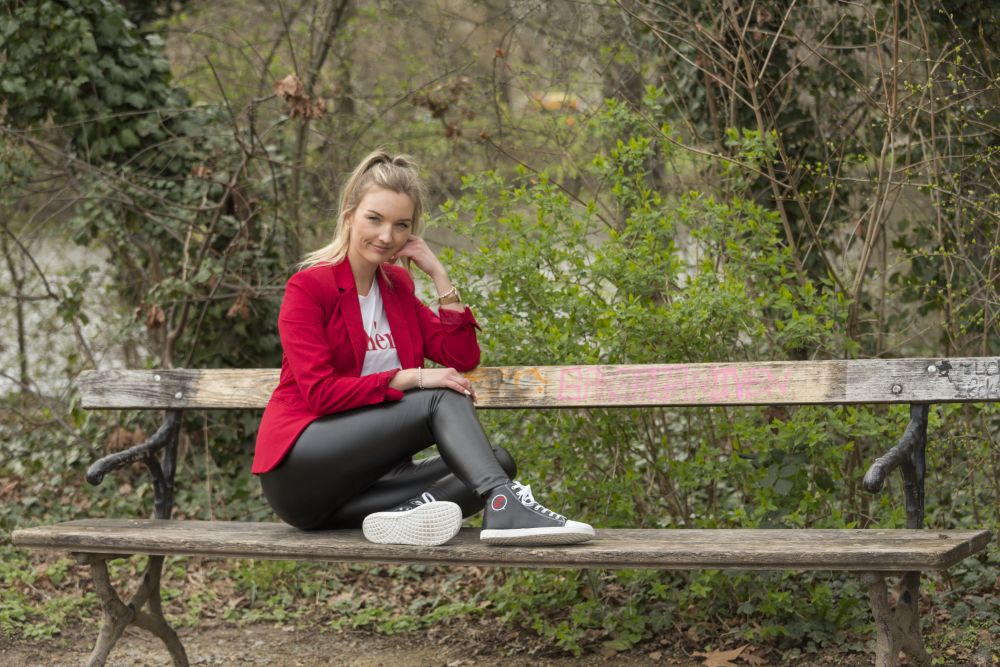 Eine Person in einem roten Blazer und einer schwarzen Lederhose sitzt auf einer Holzbank im Freien und trägt die stylischen ZAQQ CHUQQS Black High-Top-Sneaker mit Kautschuksohle. Sie lächelt, ihre Hand ruht auf ihrer Wange, umgeben von Grün und Bäumen.