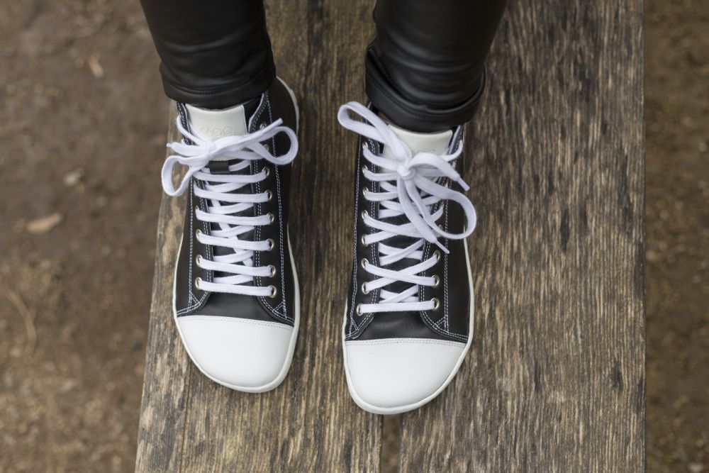 Eine Person, die schwarze High-Top-Sneakers CHUQQS von ZAQQ mit Kautschuksohle trägt, steht auf einer Holzbank. Die Schuhe haben weiße Schnürsenkel und Sohlen und die Person trägt eine schwarze Hose.