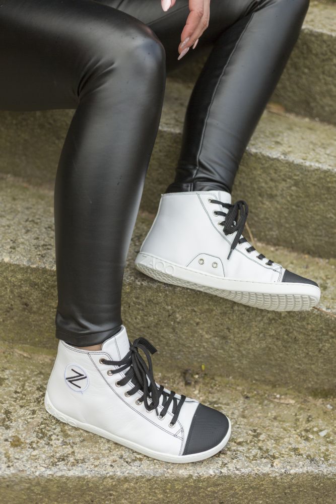 Eine Person, die schwarze Leggings und die stylischen ZAQQ CHUQQS White High-Top-Sneaker aus Nappaleder mit einem runden Emblem an der Seite und schwarzen Schnürsenkeln trägt, sitzt auf Steinstufen im Freien.