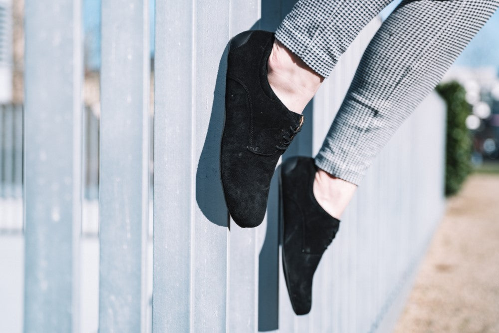 Eine Person in schwarzen IQON Veloursschuhen von ZAQQ und karierten Hosen steht auf einem Zaun im Freien. Das Velourleder der Schuhe harmoniert mit dem leicht verschwommenen Hintergrund, auf dem Gras und Bäume zu sehen sind.