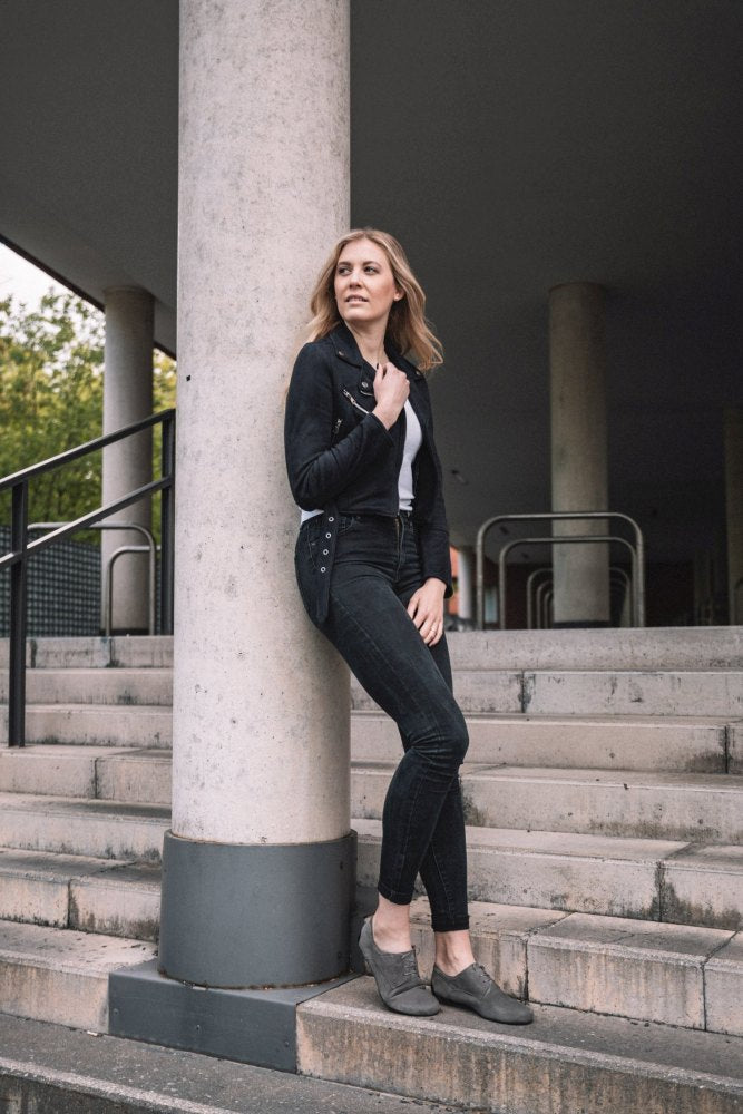 Eine Frau in einer grauen IQON Velours-Jacke und Jeans von ZAQQ lehnt an einer Säule auf den Stufen und verkörpert damit perfekt den Business-Modell-Stil. Mit einer Hand in der Tasche und der anderen in ihrem Haar strahlt sie Selbstvertrauen aus. Der Hintergrund aus Säulen und dezentem Grün rundet die Szene ab.