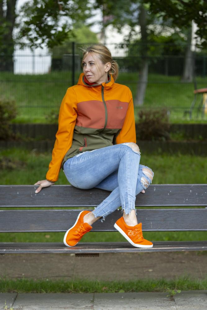 In ihren LIQE Orange Sneakers von ZAQQ entspannt eine Frau in einer orangefarbenen Jacke und zerrissenen Jeans auf einer Parkbank und genießt das angenehme Barfußgefühl. Ihre Haare sind zurückgebunden und die friedliche Umgebung wird durch grüne Bäume und einen Zaun noch verstärkt.