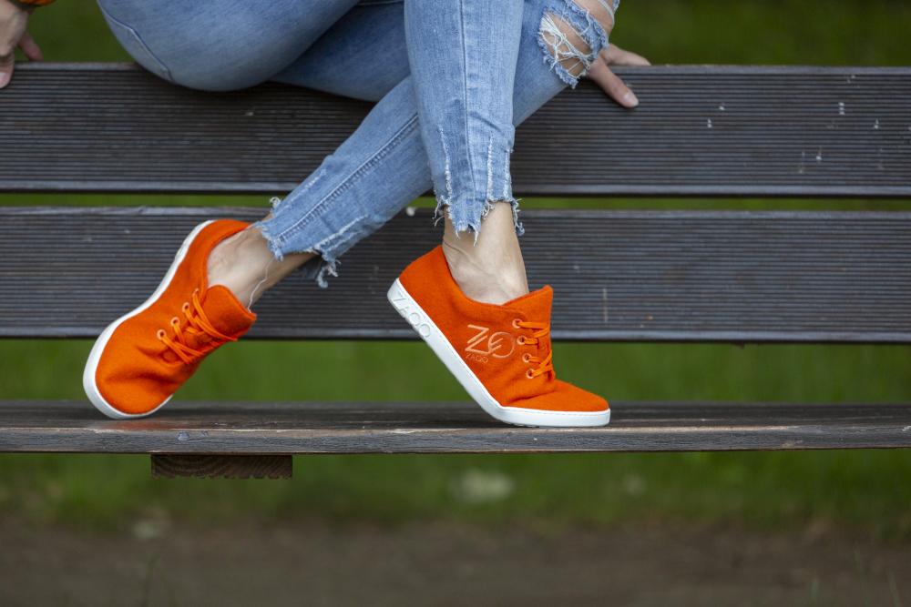 Eine Person, die die für ihr Barfußgefühl bekannten LIQE Orange-Sneaker von ZAQQ trägt, sitzt auf einer Holzbank. Sie trägt zerrissene Bluejeans vor einer grasbewachsenen Umgebung. Mit übereinandergeschlagenen Beinen genießt die Person den natürlichen Komfort, der sie umgibt.