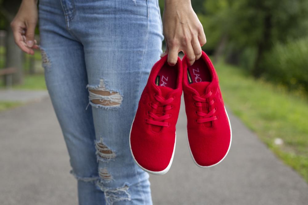 In einer Hand hält eine Person ein Paar LIQE Race-Sneaker in leuchtendem Rot mit Barfußsohlen-Technologie von ZAQQ und trägt zerrissene Bluejeans. Sie schlendern einen Außenweg entlang, umgeben von üppigem grünem Gras und Bäumen im Hintergrund.