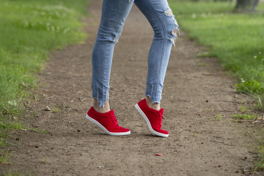 Eine Person trägt die farbenfrohen LIQE Race-Sneaker von ZAQQ mit Barfußsohle und kombiniert sie mit zerrissenen Bluejeans, während sie auf einem Feldweg durch eine Grasfläche schlendert.