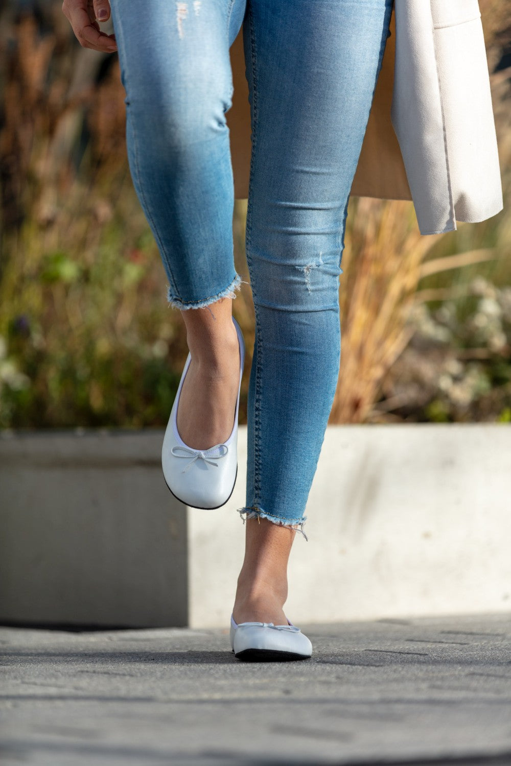 Eine Person in hellblauen Jeans und weißen ZAQQ LOOQ Ballerinas aus Nappaleder läuft anmutig auf einem Gehsteig. Im Hintergrund sind verschwommenes Grün und ein Betonpflanzgefäß zu sehen.