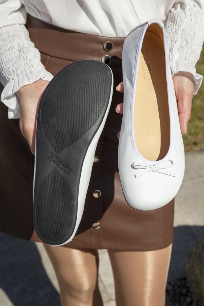 Eine Person hält elegant ein Paar LOOQ White Ballerinas von ZAQQ mit zarten Schleifen an der Oberseite. Ein Schuh zeigt seine glatte schwarze Sohle, während der andere sein raffiniertes Obermaterial aus Nappaleder hervorhebt. In einer weißen Bluse und einem braunen Rock mit Knopfdetails gekleidet, verkörpert die Person die Anmut einer Ballerina.