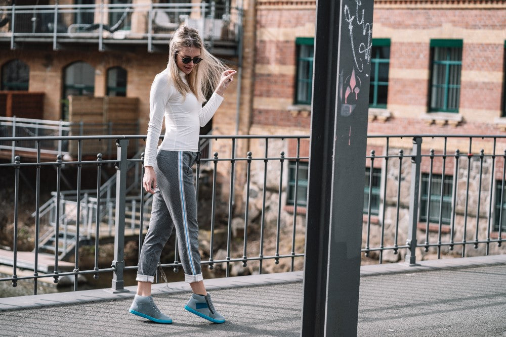 Eine Frau schlendert in LUQQ Turquoise Sneakers von ZAQQ, kombiniert mit einem weißen Langarmshirt, karierten Hosen und einer Sonnenbrille über eine Brücke. Hinter ihr ist ein mit Bogenfenstern geschmücktes Backsteingebäude zu sehen, während im Vordergrund ein mit Graffiti besprühter Metallpfosten steht.