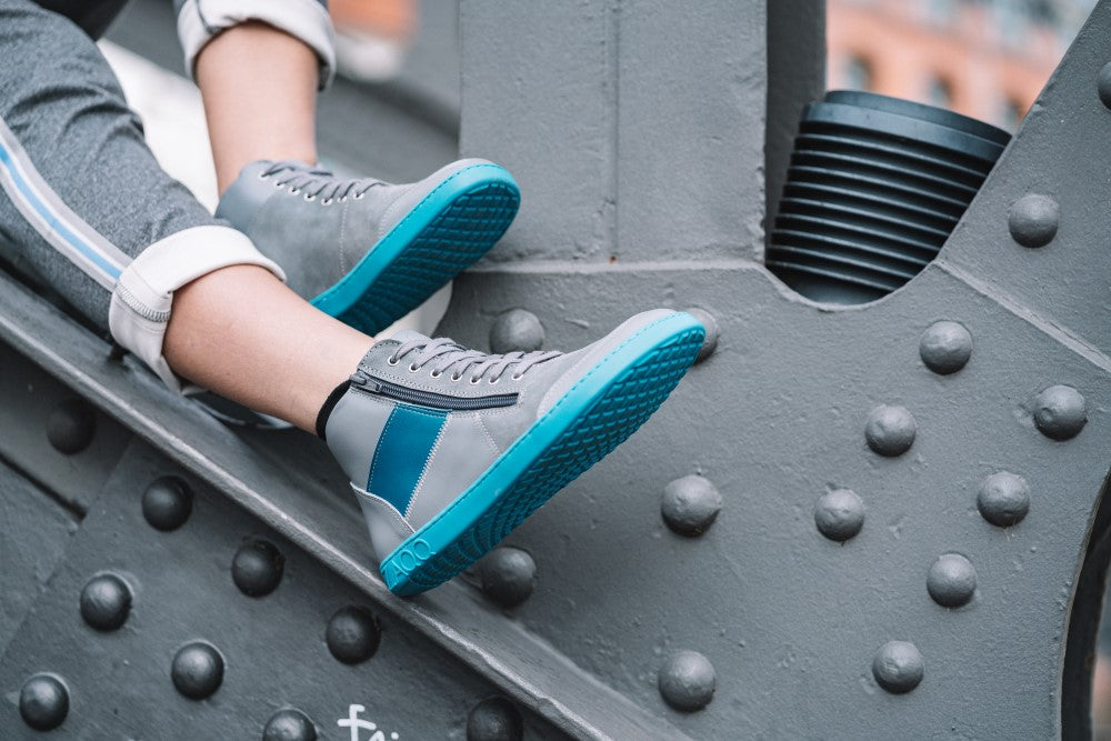 Eine Person trägt LUQQ Turquoise Sneakers von ZAQQ, ein grau-blaues Design mit hellen Kautschuksohlen, und sitzt auf einer großen Metallkonstruktion mit Nieten. Der Hintergrund zeigt eine verschwommene urbane Kulisse, die den Kontrast zwischen den Sneakers und ihrer industriellen Umgebung hervorhebt.