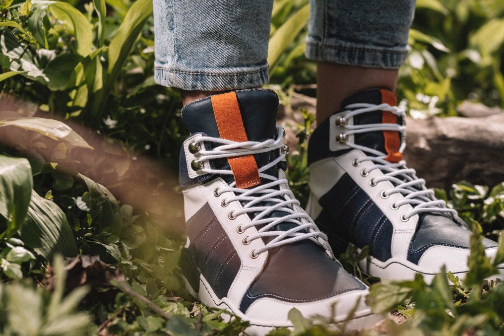 Eine Person, die die schicken MONTBLANQ Blue White Waterproof High-Top-Sneaker von ZAQQ trägt, steht auf Gras und unterstreicht die Vielseitigkeit ihres Designs. Hochgekrempelte Jeans werten das Outfit auf, während grüne Blätter und ein Baumstamm einen rustikalen Hintergrund bilden.