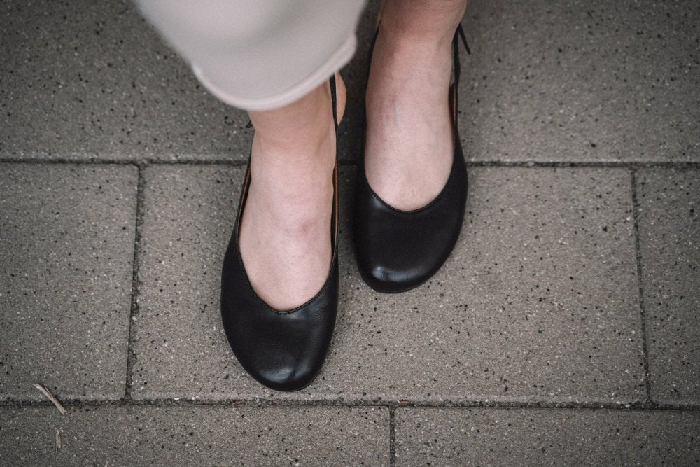 Nahaufnahme einer Person, die schwarze Ballerinas im ZAQQ PINQ-Stil auf einem gefliesten Bürgersteig trägt. Die Person trägt helle Hosen und die rechteckigen Fliesen sind grau mit sichtbaren Fugenlinien und bieten den Komfort eines Barfußschuhs.