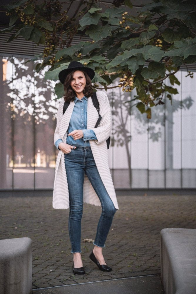 Eine Person mit breitkrempigem Hut, langer weißer Strickjacke, Jeanshemd, Jeans und eleganten dunkelbraunen QARMA-Schuhen von ZAQQ steht lächelnd unter einem Baum mit grünen Blättern und einem modernen Gebäude im Hintergrund.