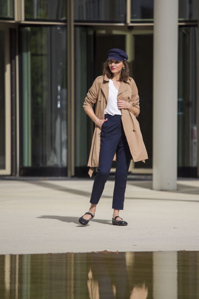 Eine Frau in hellbraunem Trenchcoat, schwarzer Kappe, Sonnenbrille, weißem Oberteil und marineblauer Hose steht selbstbewusst mit einer Hand in der Tasche da. Ihre DIEQO Nappa Black-Schuhe von ZAQQ verleihen ihr einen Hauch natürlicher Eleganz, während sie an einem sonnigen Tag im Freien posiert und ihr Spiegelbild sich klar auf der glatten Oberfläche des Gebäudes hinter ihr spiegelt.