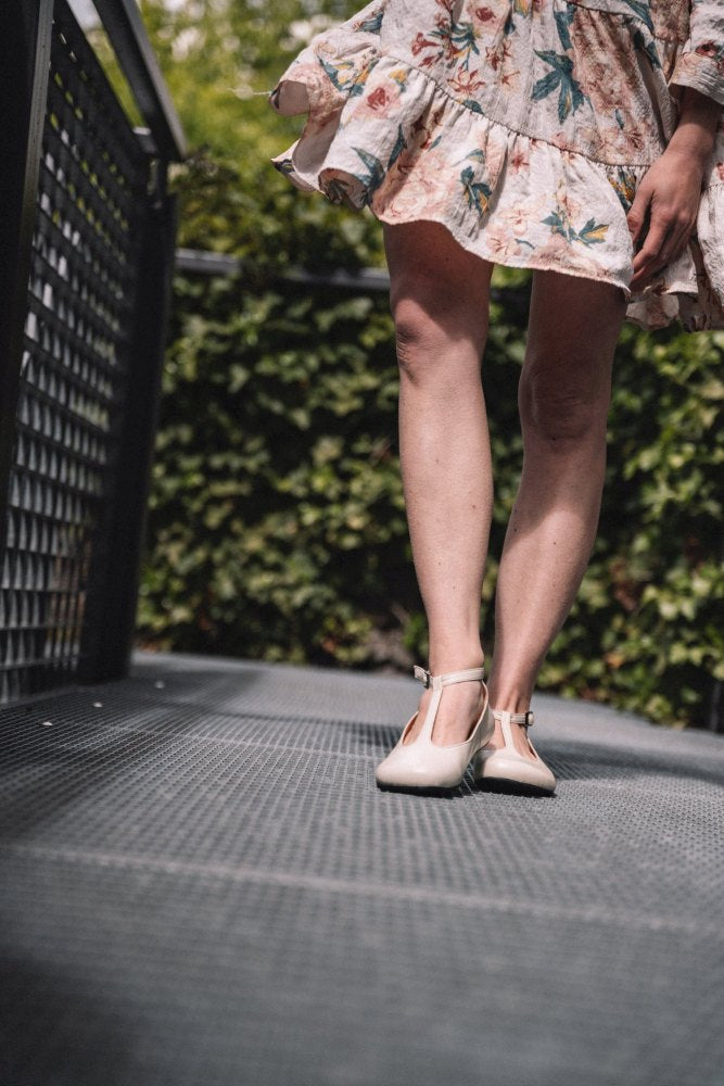 Eine Person in einem geblümten Kleid und beigen ZAQQ QITE-Schuhen steht auf einem Metallsteg, im Hintergrund ist üppiges Grün zu sehen.