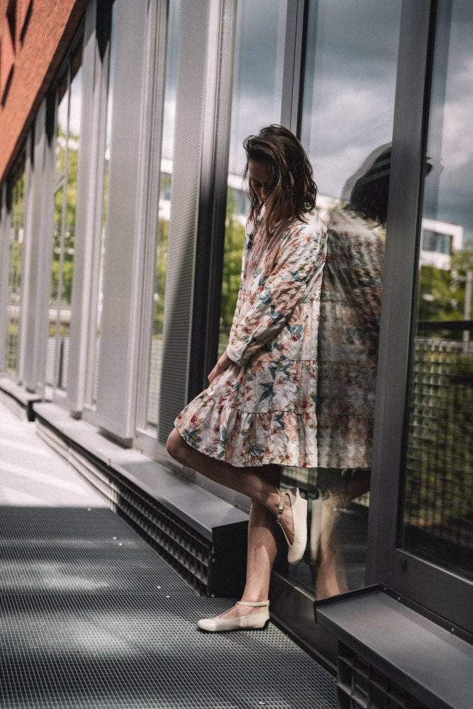 Eine Person in einem geblümten Kleid und den Ballerinas QITE Beige von ZAQQ lehnt an einer reflektierenden Glaswand auf einem Außenweg. Die Szene wird durch natürliches Licht beleuchtet und durch Grün im Hintergrund ergänzt.