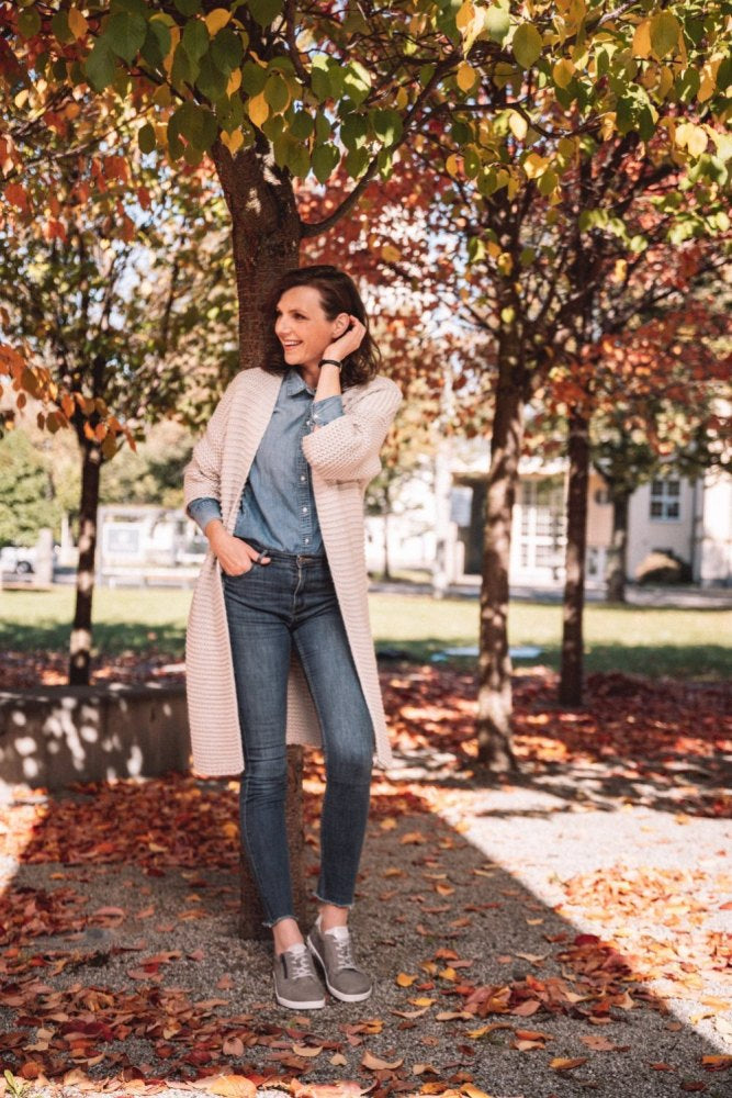 Eine Person steht unter einem Baum mit buntem Herbstlaub. Sie trägt eine leichte Strickjacke, ein Jeanshemd und Jeans dazu das kuschelige ZAQQ QOMFY Wide Grey. Sie lächelt, schaut zur Seite und hält eine Hand vor ihr Gesicht. Der Boden ist mit gefallenen Blättern bedeckt.