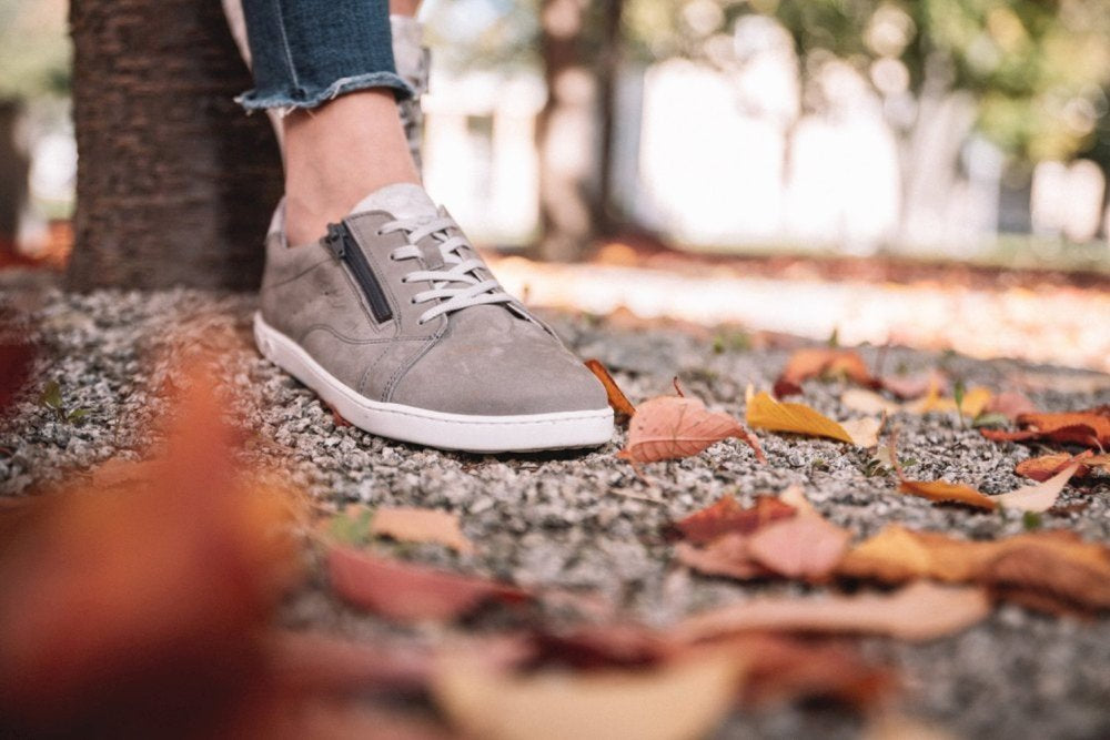 Nahaufnahme einer Person, die QOMFY Wide Grey Sneakers von ZAQQ und verkürzte Jeans trägt und auf einem Kiesweg steht. Herbstblätter liegen verstreut auf dem Boden, mit einem verschwommenen Hintergrund aus Bäumen. Der Fokus liegt auf den ZAQQ-Schuhen und Blättern, was eine herbstliche Kulisse suggeriert.