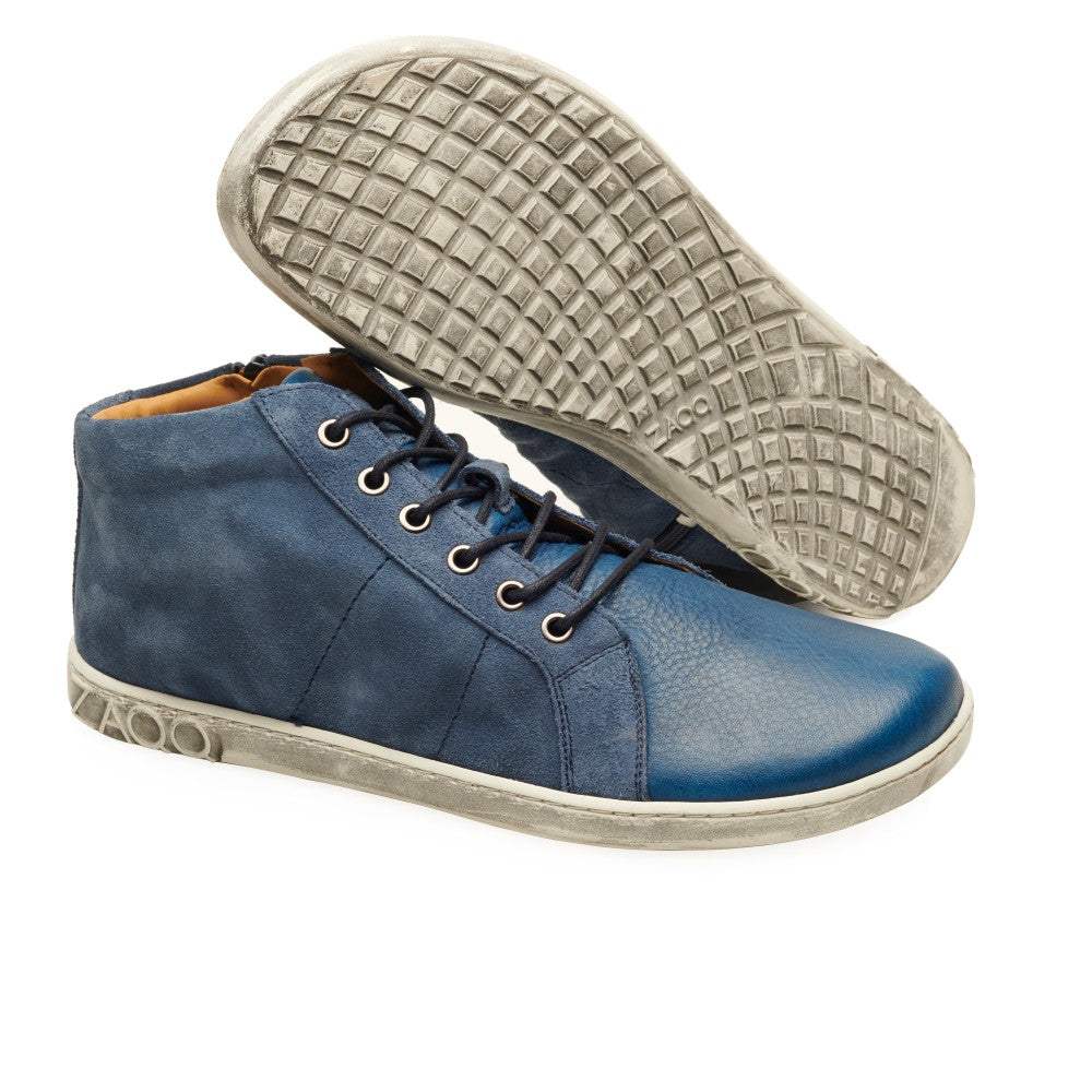 Der QORE Mid Deep Sea von ZAQQ präsentiert ein Paar blaue High-Top-Sneaker mit weißer Sohle aus Veloursleder. Ein Schuh wird aufrecht präsentiert, während der andere auf der Seite liegt, wodurch das detaillierte strukturierte Sohlendesign dieser blauen Sneaker hervorgehoben wird.