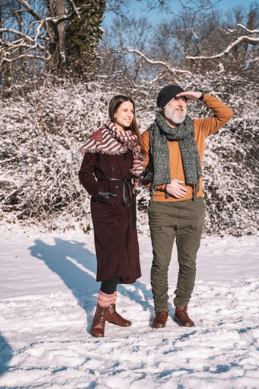 Ein Mann und eine Frau stehen in einer verschneiten Landschaft. Die Frau trägt einen langen Mantel und einen Schal, während der Mann eine Beanie, einen orangefarbenen Pullover und QUINTIC Winter Vegan Nut Boots von ZAQQ trägt und seine Augen mit der Hand abschirmt. Den Hintergrund bilden schneebedeckte Bäume mit Ästen.