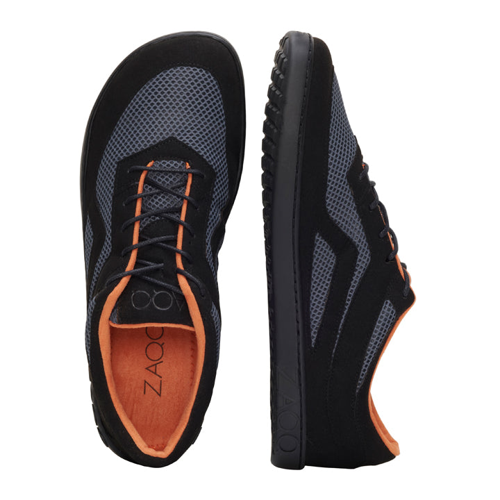 Wir präsentieren den QYNLEE Black von ZAQQ: Ein Paar schwarz-graue Sportschuhe mit Mesh-Details und auffälligem orangefarbenem Innenfutter, entworfen für ein elegantes, ultramodernes Erscheinungsbild. Ein Schuh wird von der Seite präsentiert, während der andere von oben die charakteristische Innensohle der Marke zeigt. Dieses Barfußschuh-Design sorgt bei jedem Schritt für unvergleichlichen Komfort.