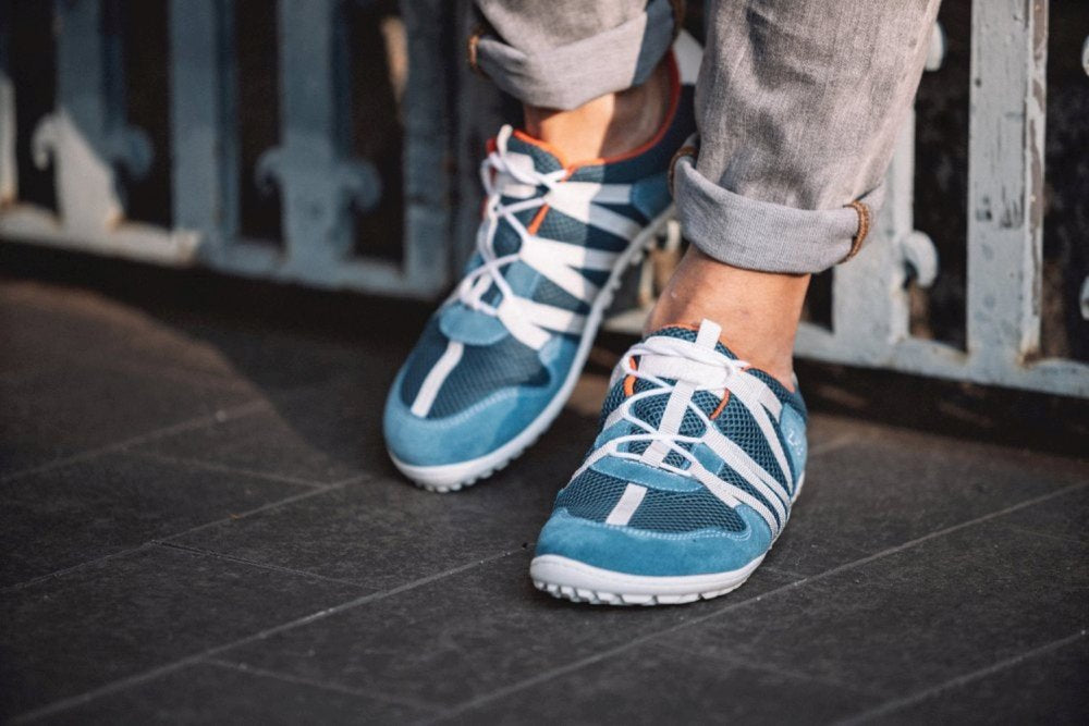 Eine Person, die RIQ Blue-Sneakers mit weißen Schnürsenkeln von ZAQQ trägt, steht auf einer dunklen Fläche, kombiniert mit hochgekrempelten grauen Jeans. Diese blau-weißen Schuhe bestehen aus atmungsaktivem Netzgewebe und bieten so mehr Komfort. Im Hintergrund ist ein dekoratives Metallgeländer zu sehen.