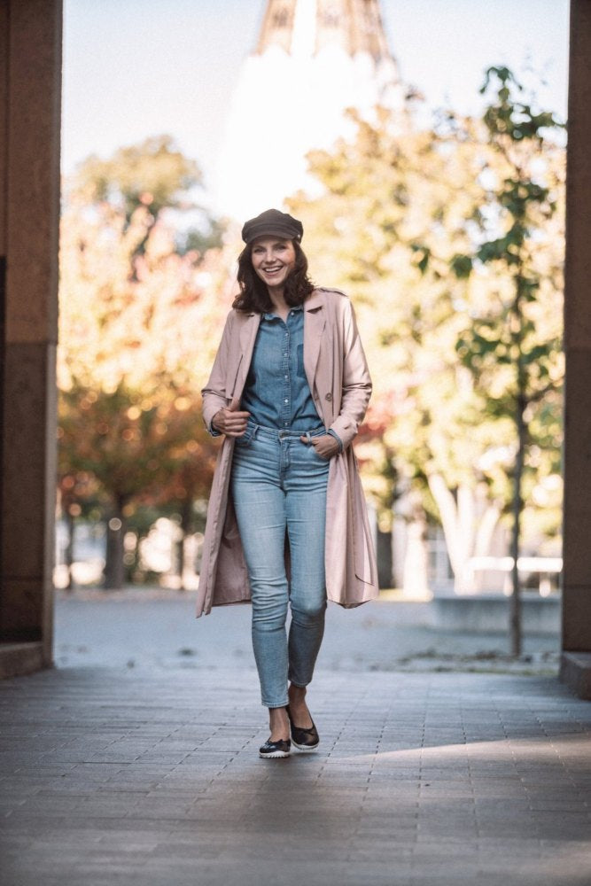 Eine Person lächelt, während sie einen sonnenbeschienenen Weg im Freien entlangspaziert. Sie trägt einen rosa Mantel, ein Jeanshemd, Jeans und eine dunkle Mütze. Um ihr Outfit zu vervollständigen, trägt sie die dunkelblauen SUQAR-Schuhe von ZAQQ, während Bäume mit Herbstlaub den Hintergrund säumen.