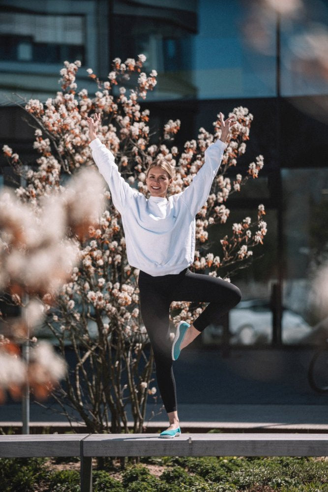 Eine Person, die einen SUQAR Heaven-Pullover von ZAQQ und schwarze Leggings trägt, balanciert auf einem Bein neben einem blühenden Baum, die Arme zu einem V erhoben, was perfekte Atmungsaktivität demonstriert. Im Hintergrund ist ein modernes Gebäude mit Glasfenstern zu sehen.