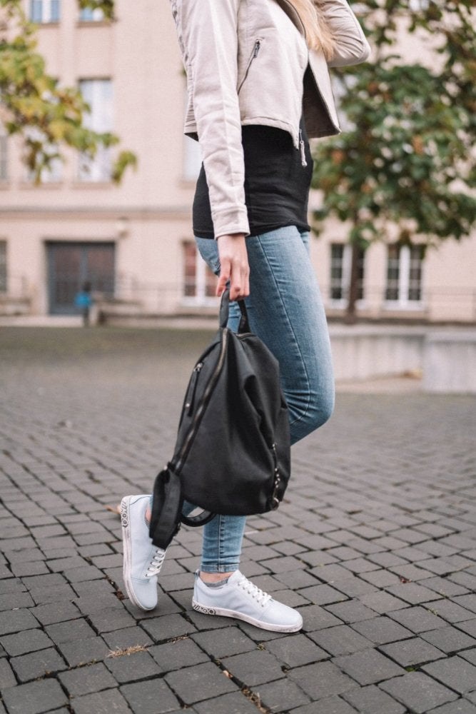 Eine Person läuft auf einem Kopfsteinpflasterweg. Sie trägt eine leichte Jacke, ein schwarzes Hemd, blaue Jeans und weiße Sneakers mit der flexiblen Sohle des ZAQQ TAQQ Nappa Lightblue und hält einen schwarzen Rucksack in der Hand. Im Hintergrund sind ein Gebäude und Bäume mit grünen Blättern zu sehen.