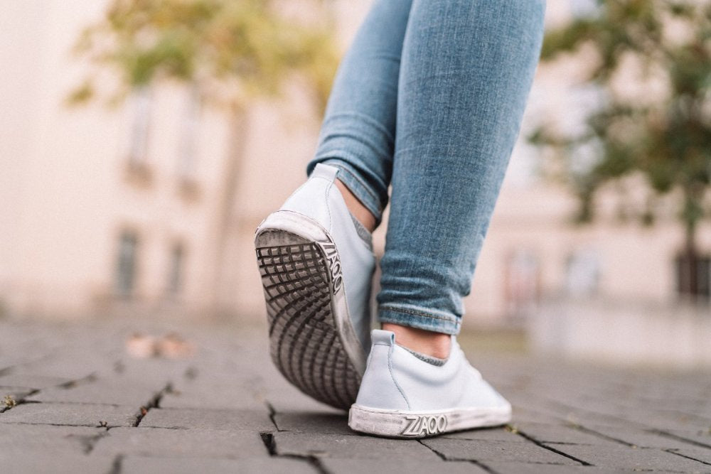 Eine Person trägt hellblaue Jeans und ein Paar TAQQ Nappa Lightblue-Sneaker von ZAQQ und läuft auf einem Kopfsteinpflasterweg. Der Fokus liegt auf ihren Füßen, die die flexible Sohle der Schuhe zeigen, mit verschwommenen Bäumen und Gebäuden im Hintergrund. Auf den Sohlen der Sneaker ist Text zu sehen.