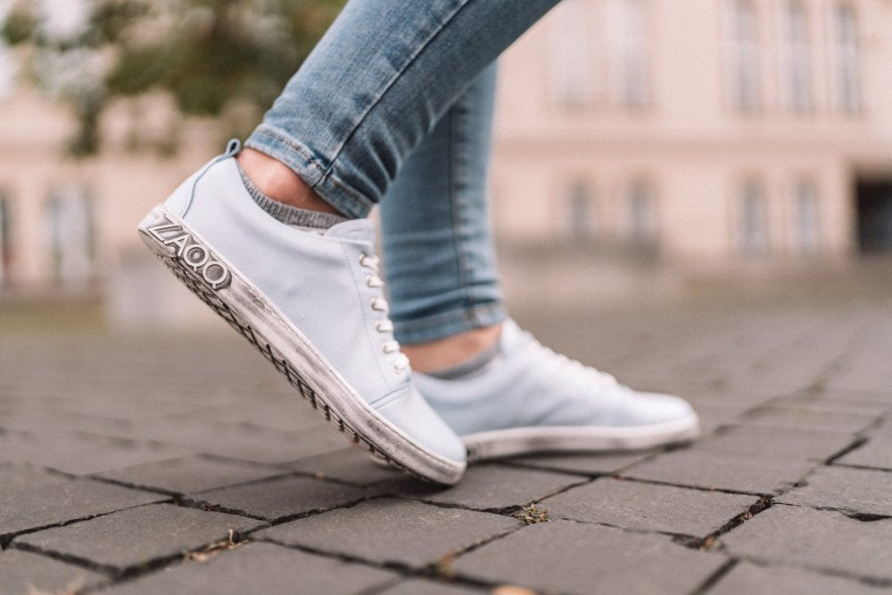 Eine Person trägt die TAQQ Nappa Lightblue Sneakers von ZAQQ und Blue Jeans und läuft auf einem Kopfsteinpflasterweg. Im Hintergrund ist ein unscharfes beiges Gebäude zu sehen.