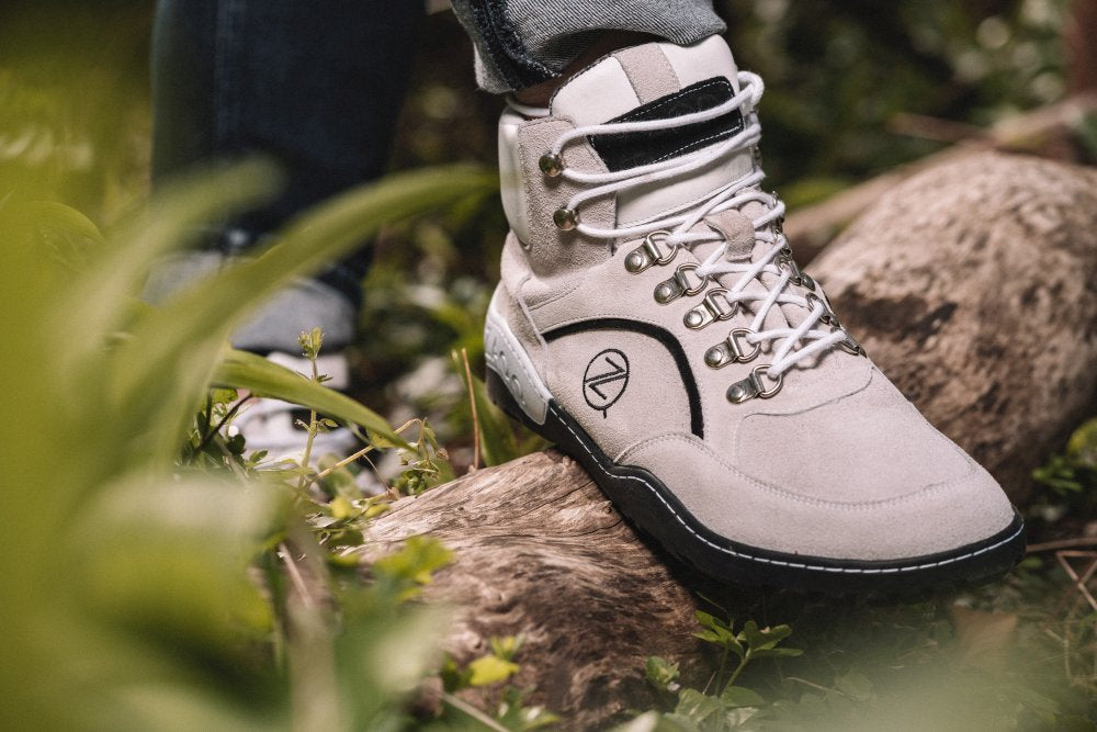 Nahaufnahme der TERRAQ Beige Waterproof Wanderschuhe von ZAQQ mit weißen Schnürsenkeln und schwarzen Akzenten, getragen von einer Person, die auf einem Baumstamm steht, umgeben von Grün. Diese Outdoorschuhe haben ein kreisförmiges Logo an der Seite.